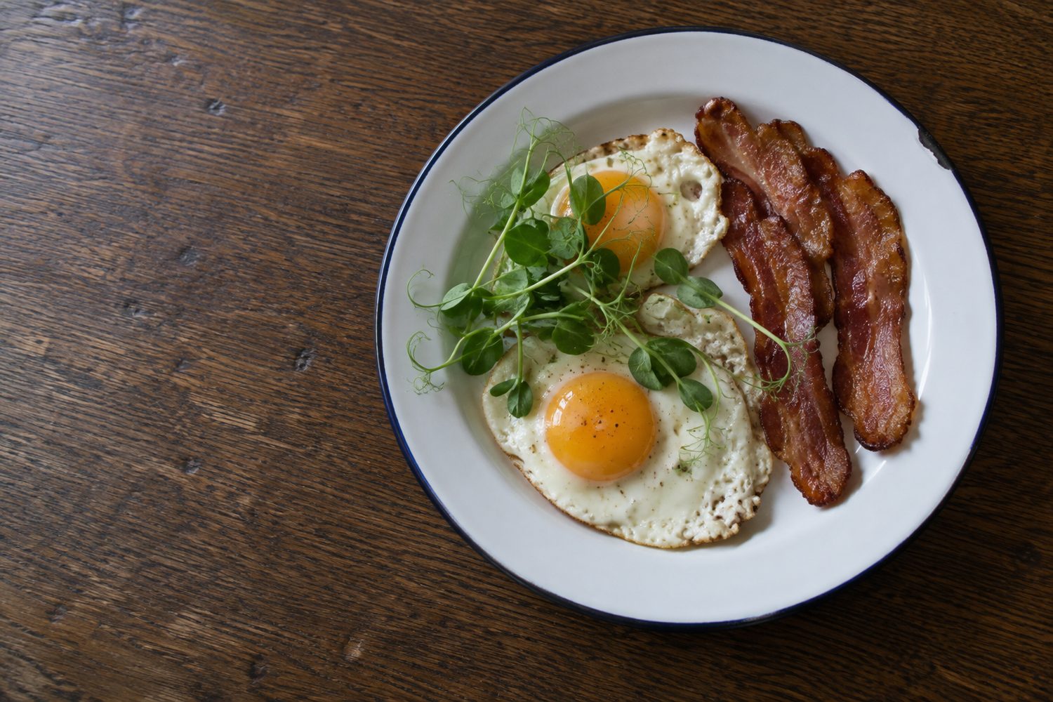 Breakfast plate of two fried eggs and crisp bacon topped with a small tangle of pea shoots on a wooden kitchen table