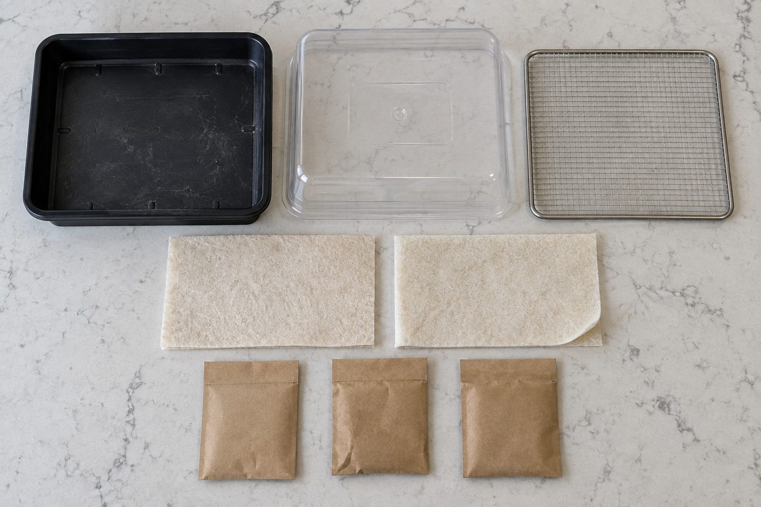 Parts of a microgreen kit laid out on a counter: a shallow tray, a clear humidity dome, a mesh insert, two thin fiber grow pads and three plain seed pouches