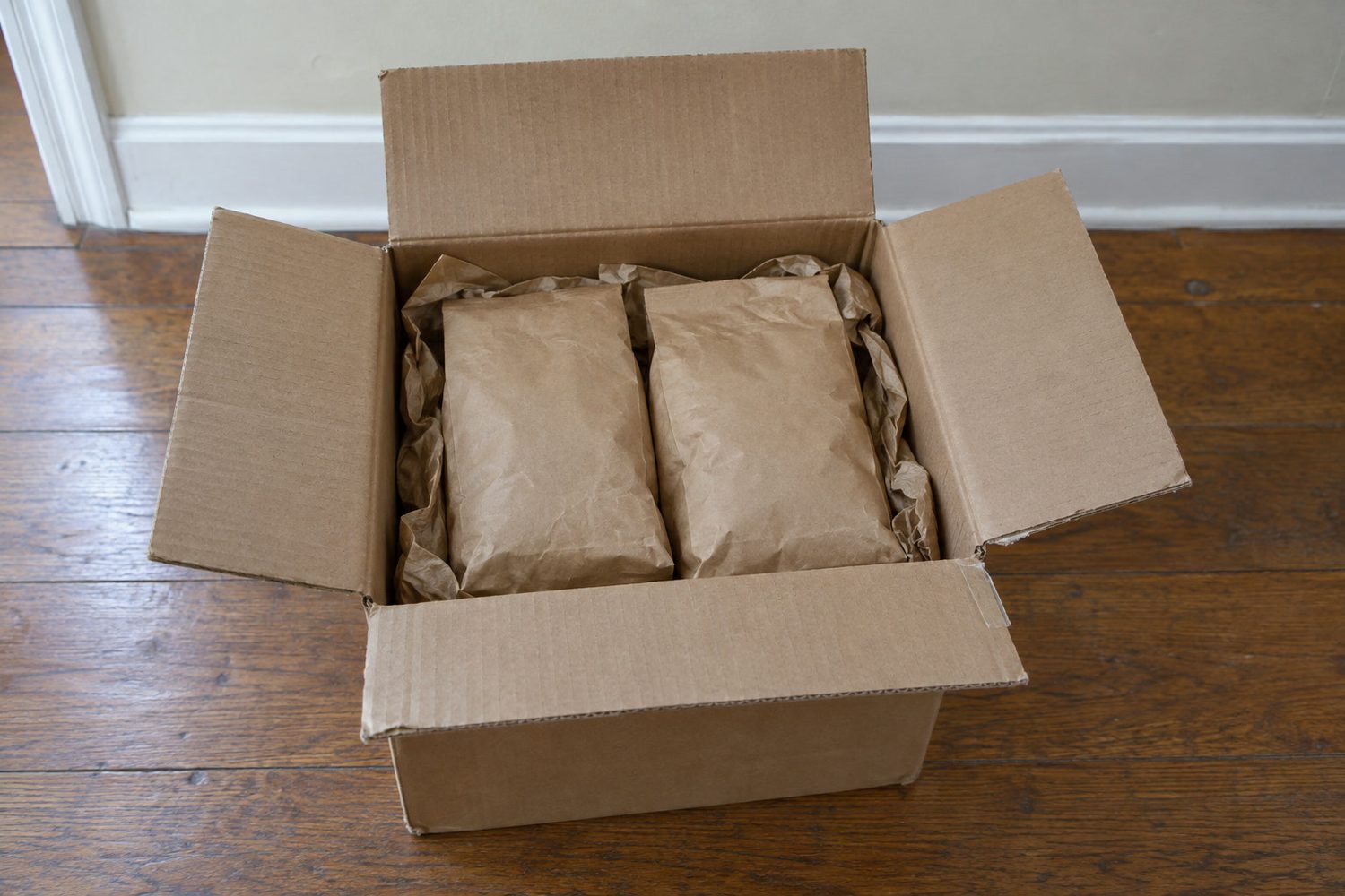 Opened plain brown cardboard shipping box on a wood hallway floor holding two plain unmarked paper seed bags nestled in crumpled brown packing paper