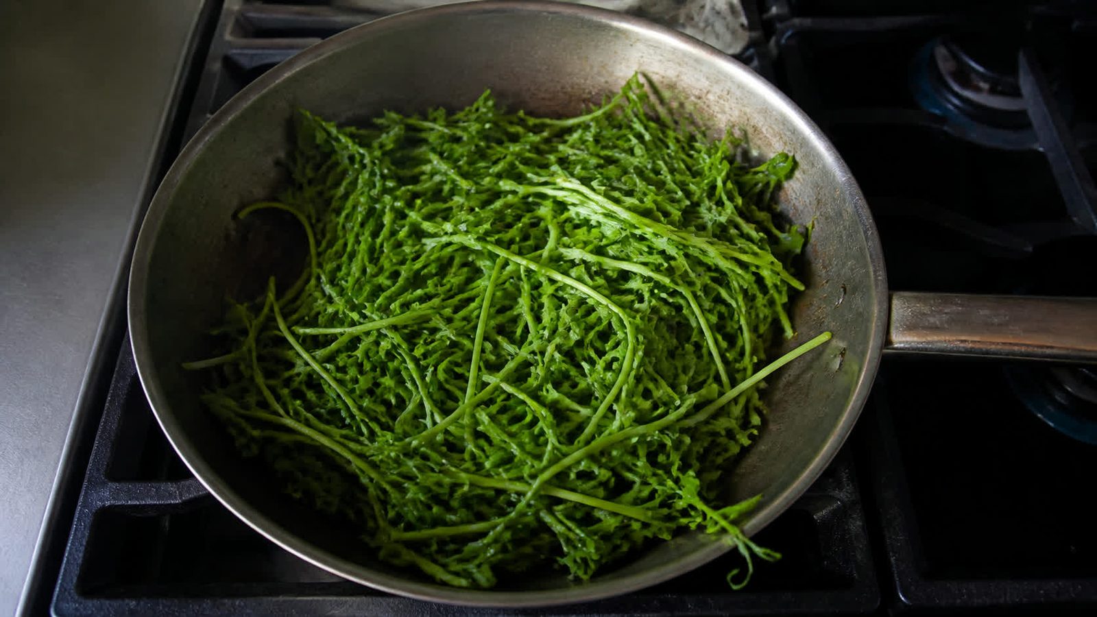 A wide skillet on a hob holding a heaped pile of pea shoots just beginning to wilt, photographed from above in flat daylight with the stems still distinct rather than collapsed