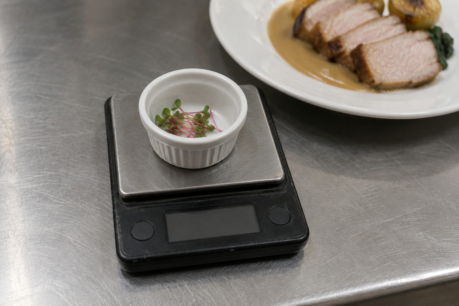 A tiny pinch of radish microgreens in a small white ramekin on a compact pocket scale with a dark blank display, next to a plated dish on a prep counter