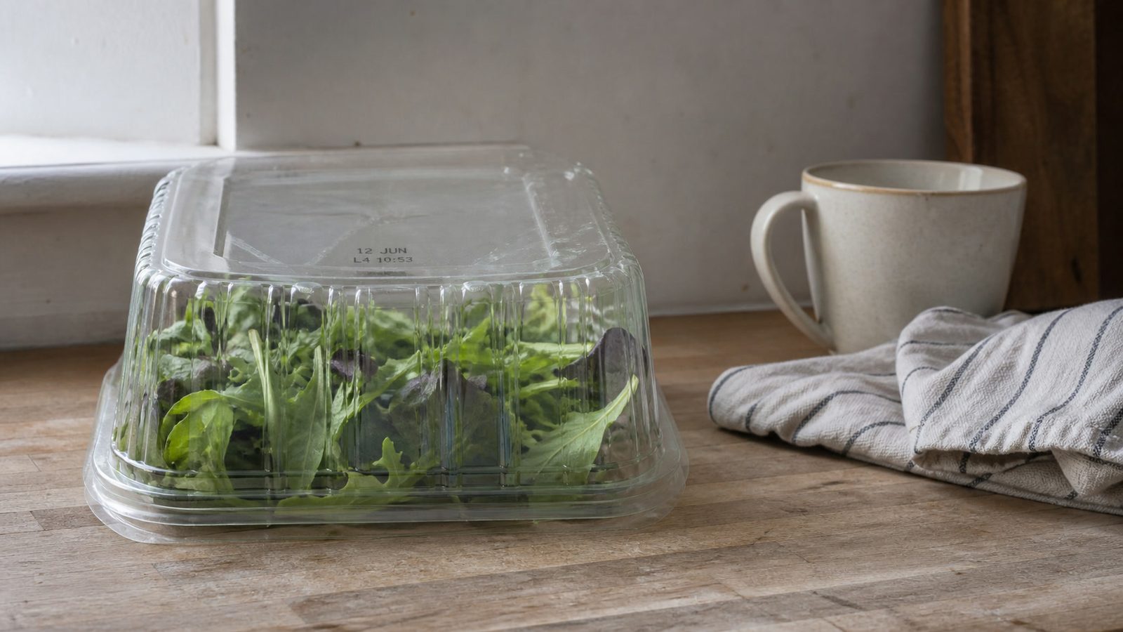 A closed plain clear clamshell of mixed salad leaves on a worn wooden counter beside a window, photographed side on at counter height in flat daylight, a short inkjet code stamped on the top of the lid reading 12 JUN and L4 10:53, with a plain ceramic mug and a folded striped tea towel to the right and no brand names or packaging artwork anywhere in frame