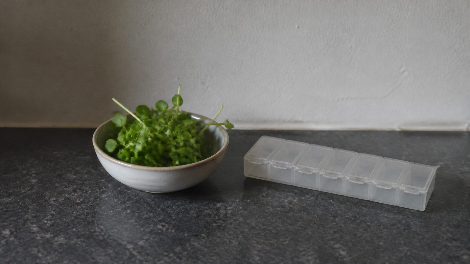 A small bowl of cut green microgreens on a kitchen counter beside a weekly pill organizer, photographed level with the counter in flat daylight
