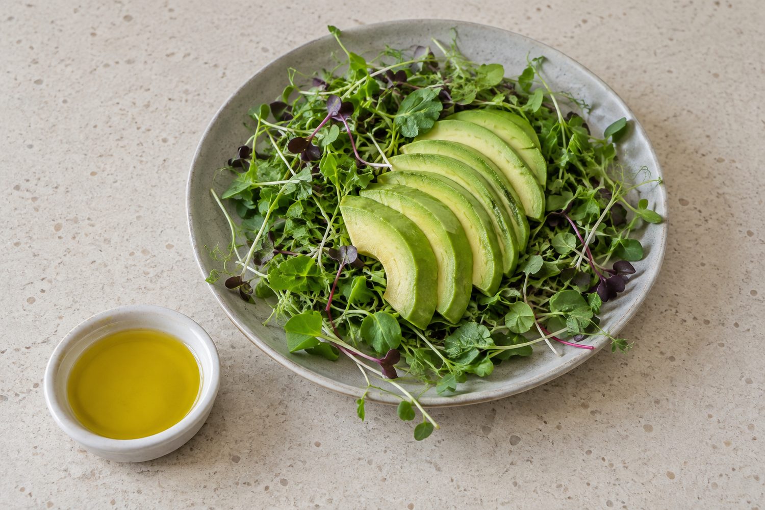 Salad of mixed microgreens topped with slices of ripe avocado beside a small dish of olive oil on a light kitchen counter