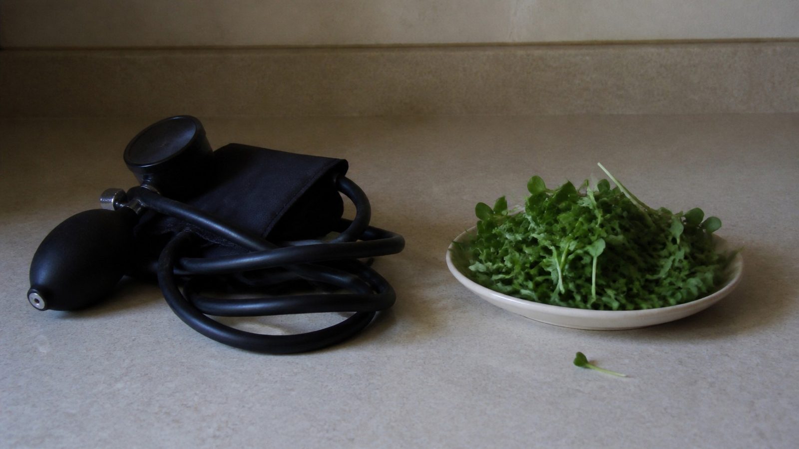 A blood pressure cuff and a small dish of cut green microgreens side by side on a plain kitchen counter, photographed level with the counter in flat daylight