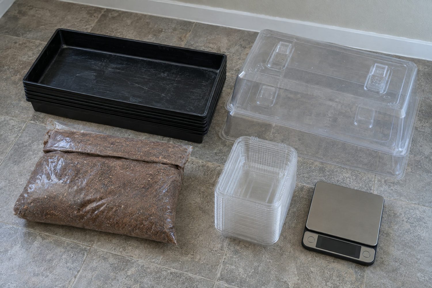 Microgreens starter equipment laid out on a floor: a stack of shallow black trays, clear humidity domes, a plain bag of growing medium, a sleeve of clear clamshells and a small scale with a dark display