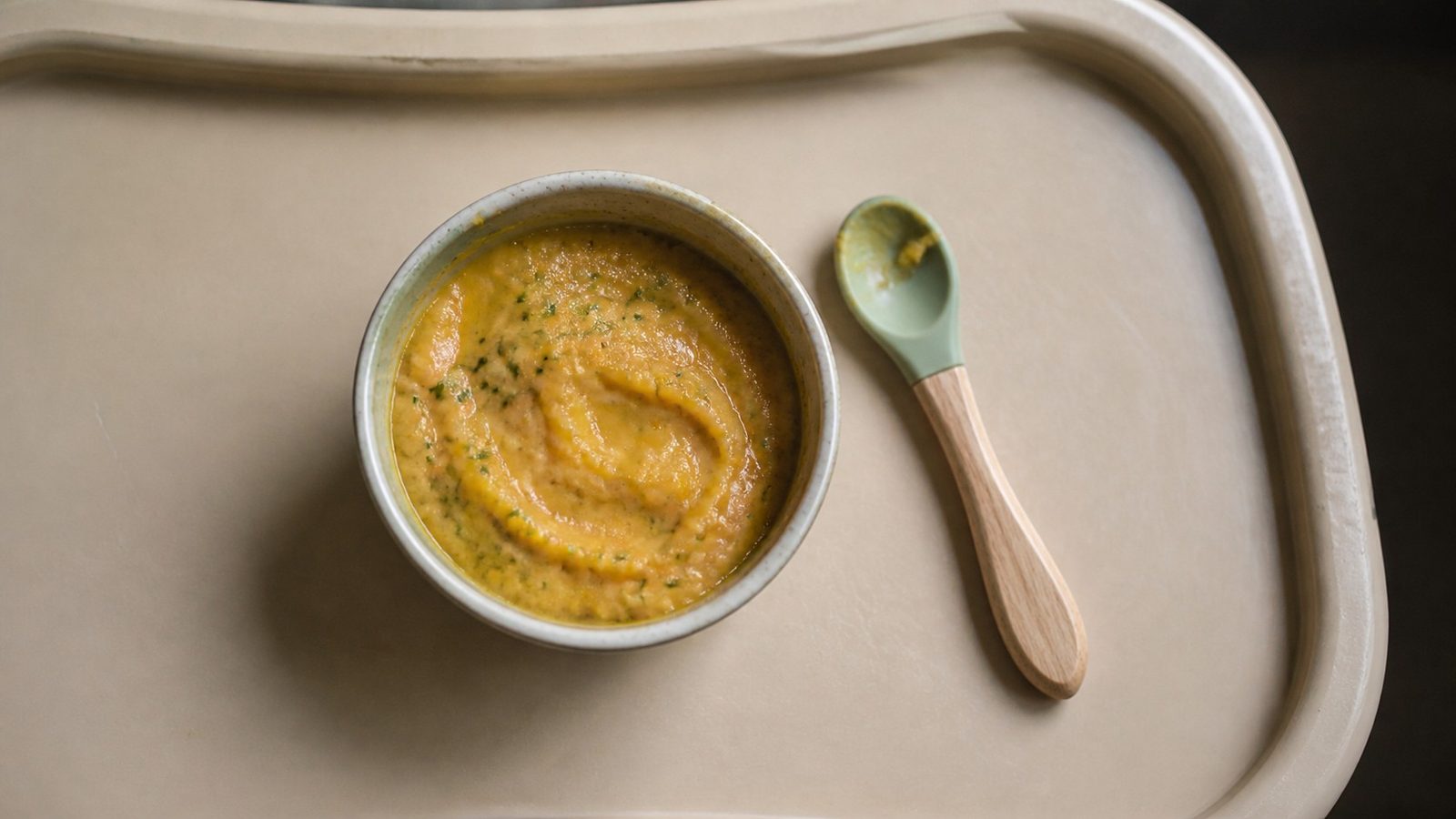A small bowl of soft cooked vegetable puree with finely minced green microgreens stirred through it, set on a high chair tray beside a short-handled weaning spoon, photographed from above in flat daylight