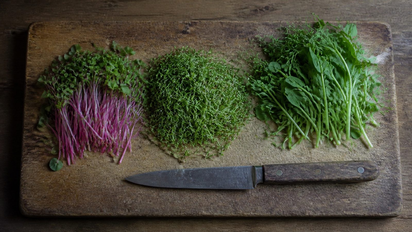 A cutting board in flat daylight with three small piles of harvested microgreens side by side, radish, broccoli and pea, photographed from just above the board with a knife resting beside them