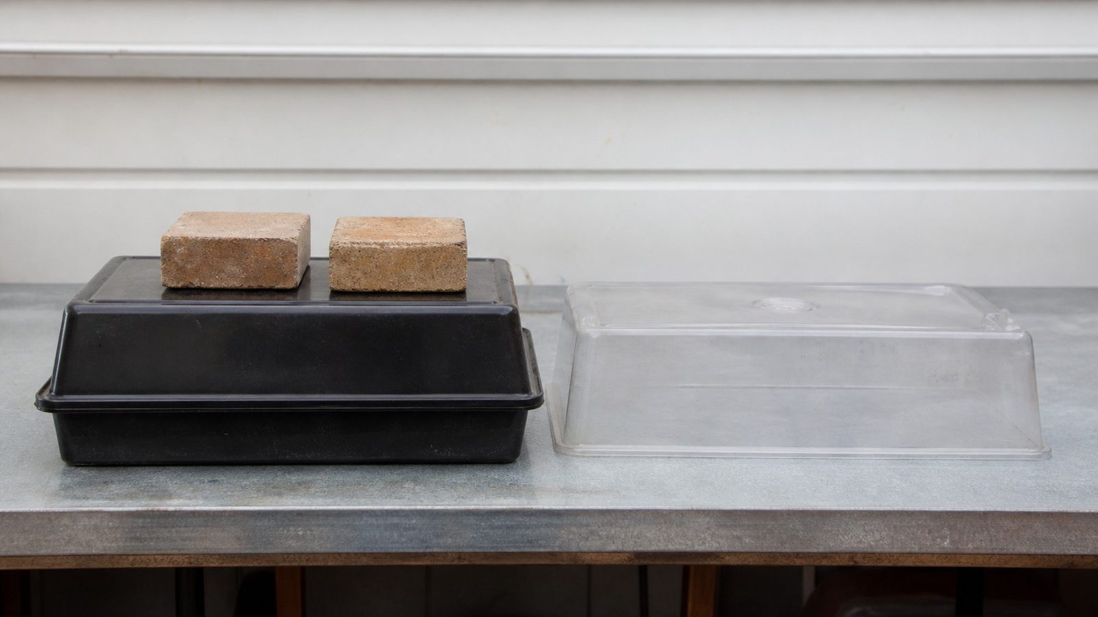 Two 1020 trays stacked on a bench, the upper one inverted over a freshly sown tray with a couple of bricks resting on top as weight, beside a clear plastic humidity dome standing unused, photographed level with the bench in flat indoor light